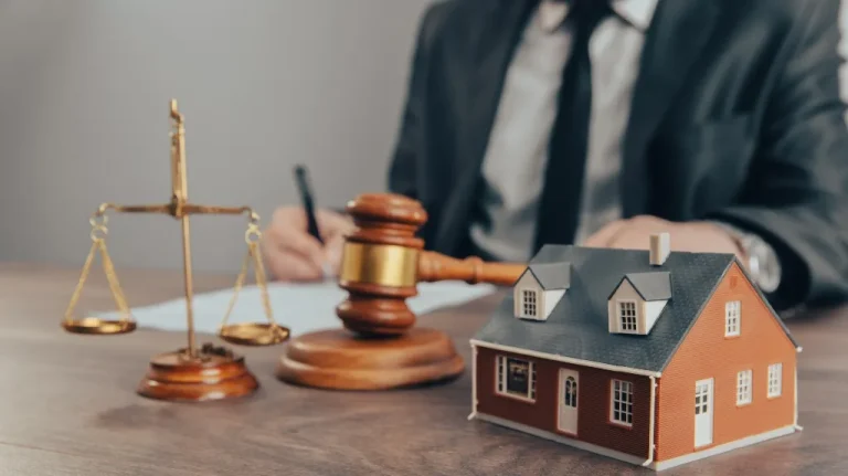 A small model house, wooden gavel, and scales of justice on a desk with a person in a suit writing in the background.