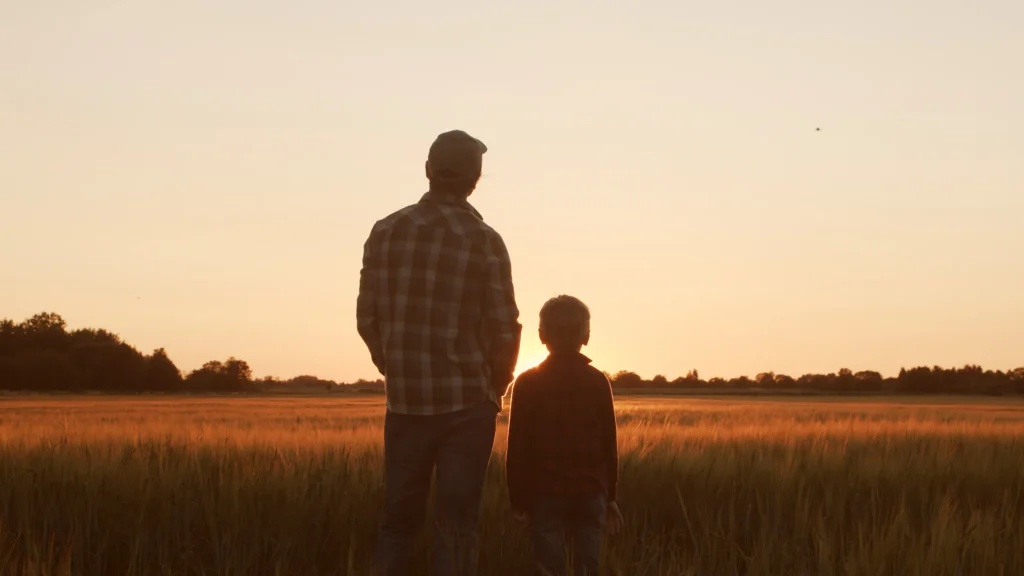 Silhouettes of an adult and child standing in a field at sunset with a clear sky.