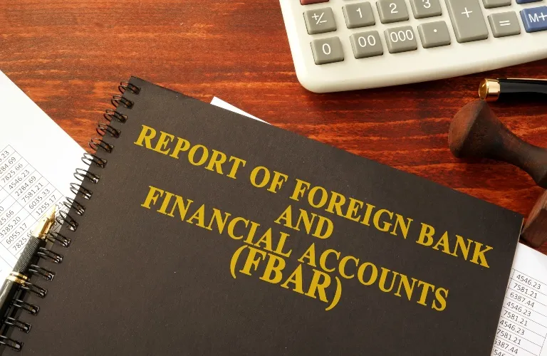 Black spiral-bound report titled "Report of Foreign Bank and Financial Accounts (FBAR)" on wooden desk with calculator, pen, and financial documents.