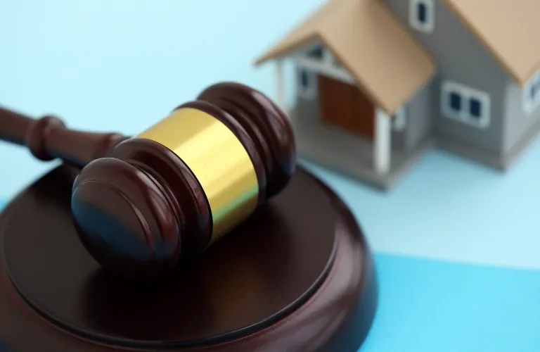 Wooden judge's gavel with gold band beside a small gray model house on blue surface.