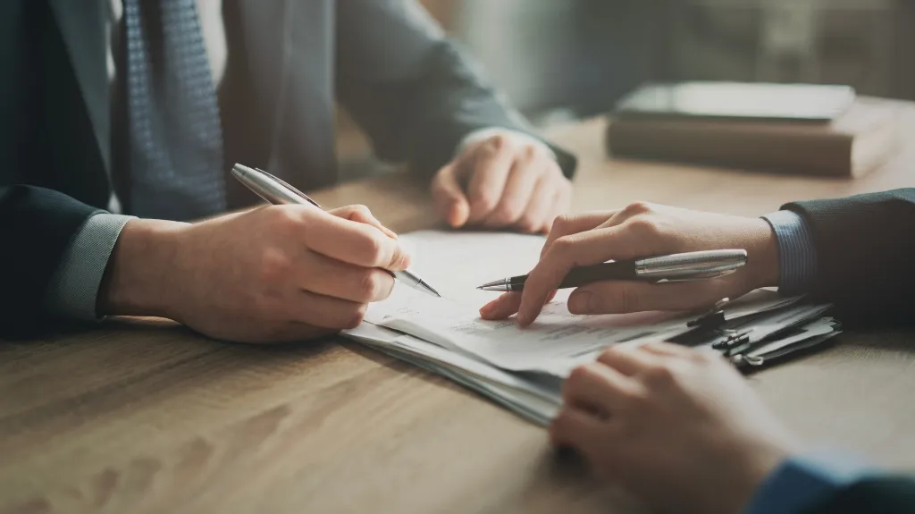 Two individuals in business attire reviewing and signing documents with pens on a wooden table.