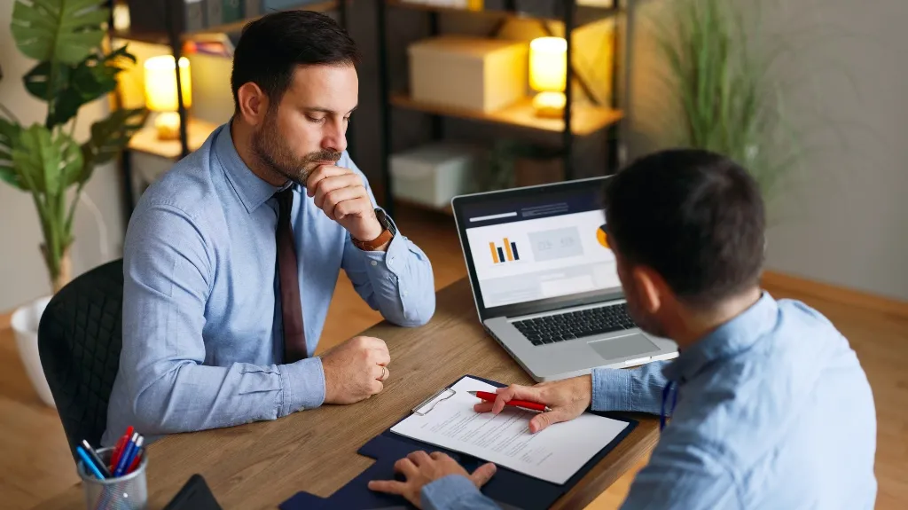 Two men in blue shirts discussing documents at an office desk with a laptop.