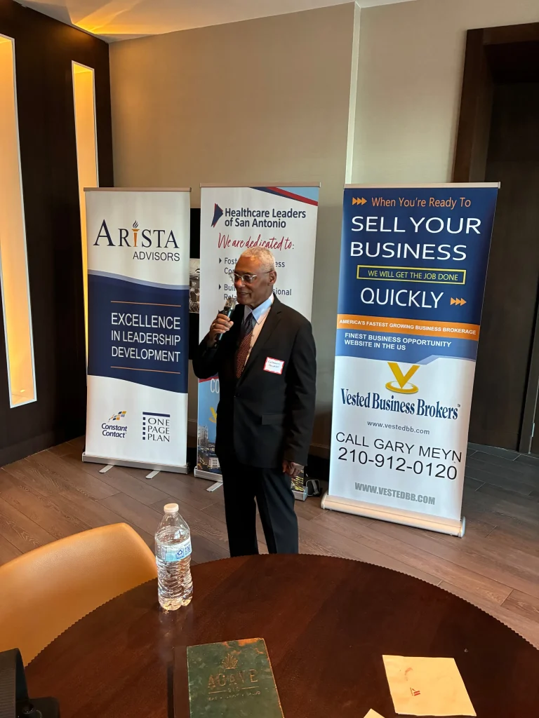 Man in dark suit speaking into microphone, standing between three business promotional banners.