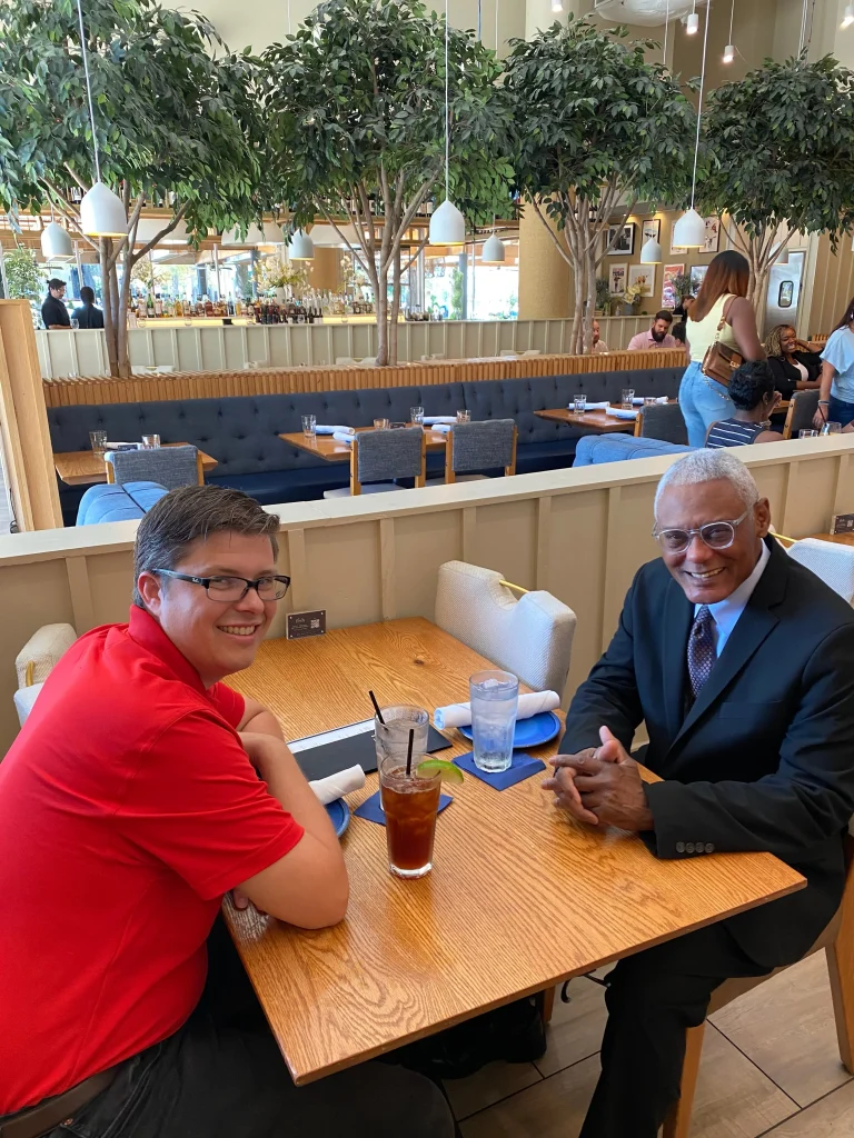 Two men smiling at a restaurant table with drinks, indoor plants, and patrons in background.