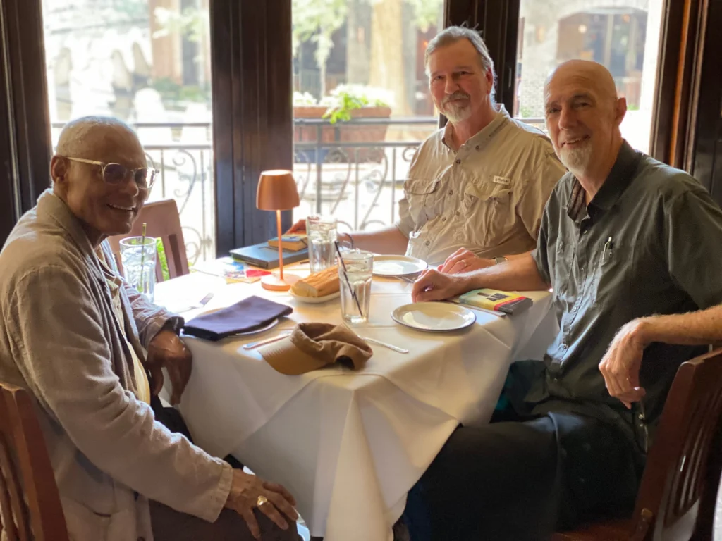 Three older men seated around a white-clothed table in a restaurant with bread, glasses, and books.