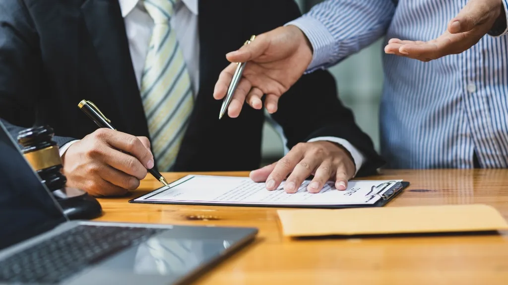 Two men in business attire reviewing and signing a legal document on a wooden table.