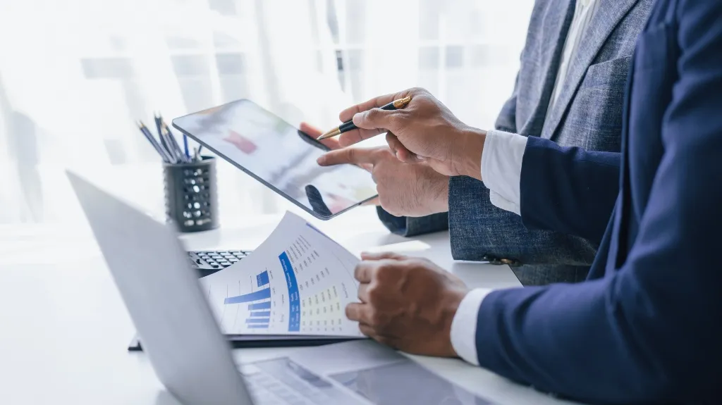 Two professionals in business attire reviewing charts and data on a tablet and printed documents.