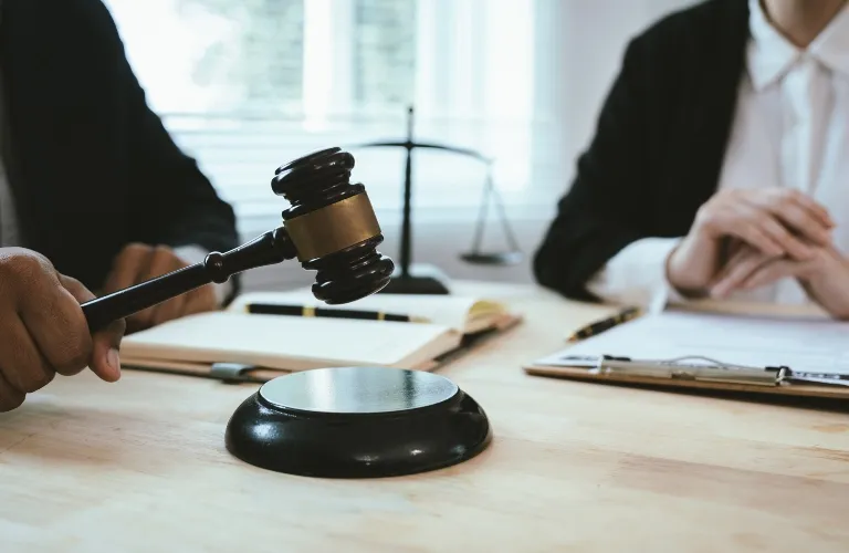 Judge's hand holding a wooden gavel above a sound block on a desk with legal documents and a balance scale in the background.