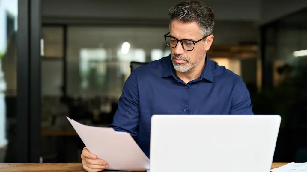 Middle-aged man with glasses and gray hair reviewing documents behind a laptop in an office.