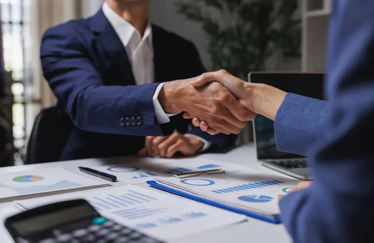 Two business professionals in suits shaking hands over a desk with financial documents and a laptop.