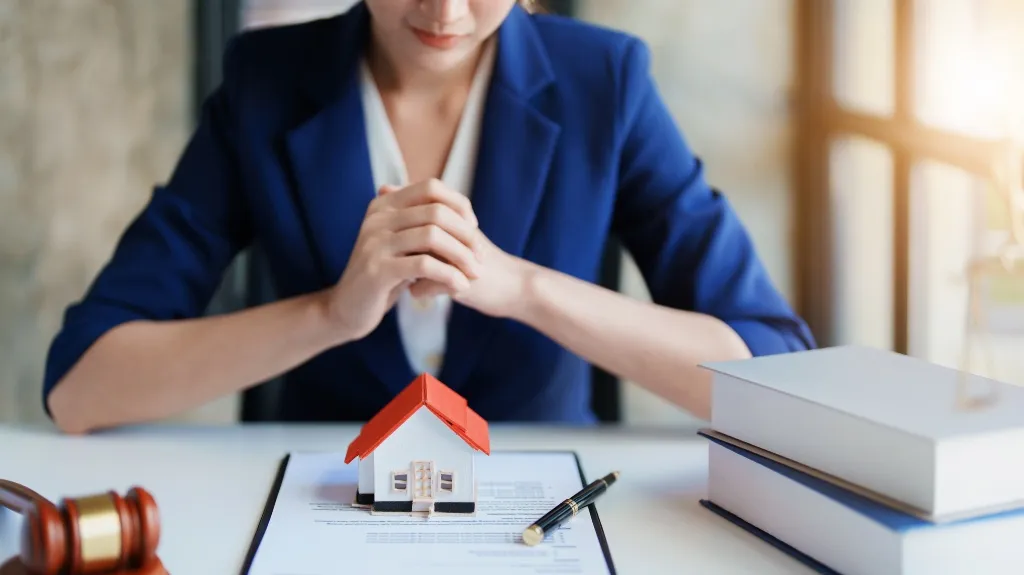 Professional woman in blue blazer with house model, legal documents, books, and gavel on desk.