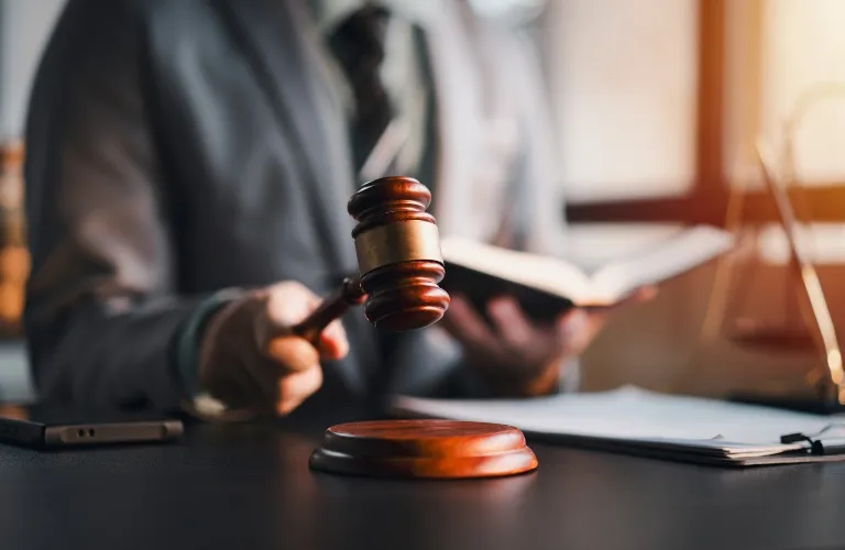 Person in suit holding wooden gavel above sound block with open book and documents on desk.