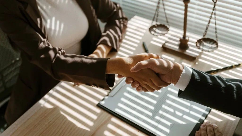 Two business professionals shaking hands over a desk with legal scales and documents.