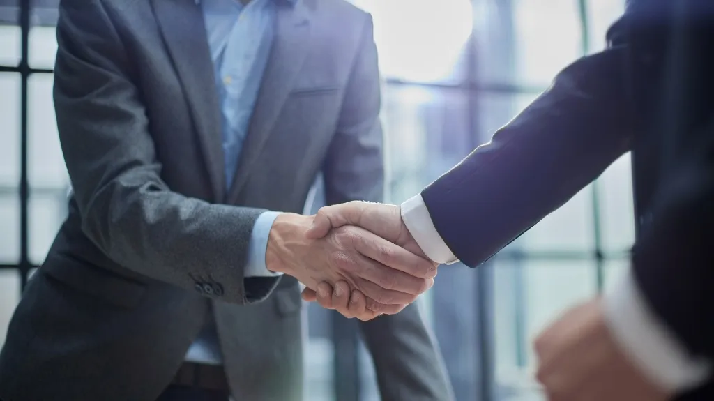 Two business professionals in suits shaking hands in a modern office setting.