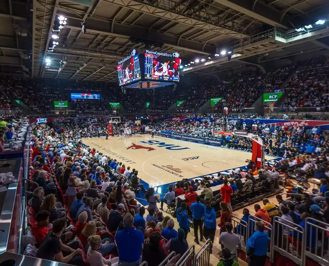 Indoor basketball arena filled with spectators, players, and large digital scoreboard.
